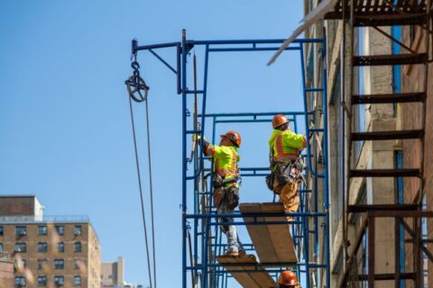 Construction workers wearing safety gear on scaffolding at height.