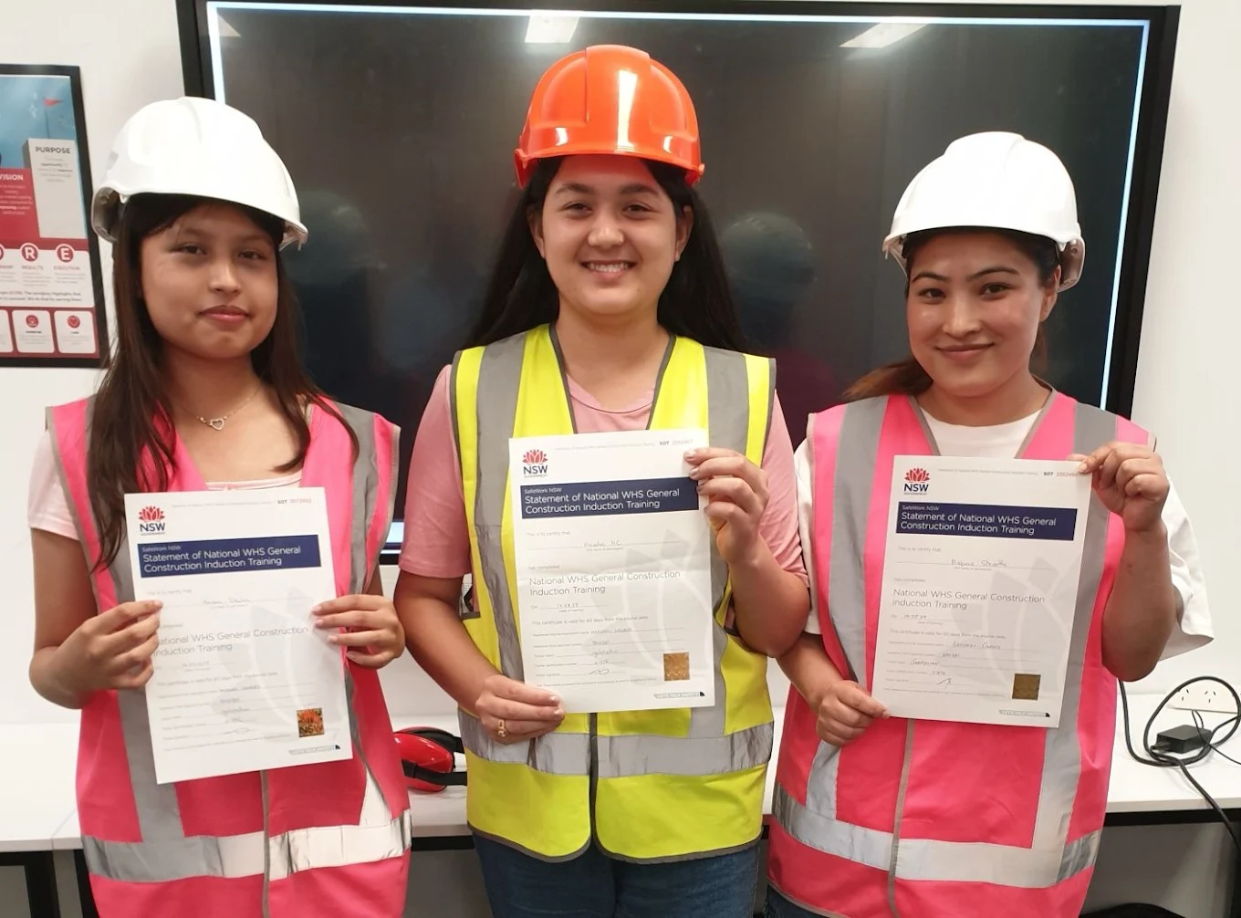 Three trainees wearing safety vests and helmets, holding White Card certificates after finishing construction induction training.